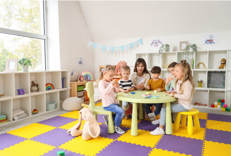 Children and a teacher sit around a lime round table doing crafts in a bright, colorful classroom with yellow and purple floor mats.